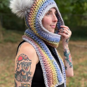 May include: A rainbow crochet hooded scarf with a fluffy pom pom. The scarf is made with a variety of colors, including pink, purple, blue, green, and yellow. The scarf is tied around the neck of a person with tattoos on their arms. The scarf has a tag that says "Of Mars".