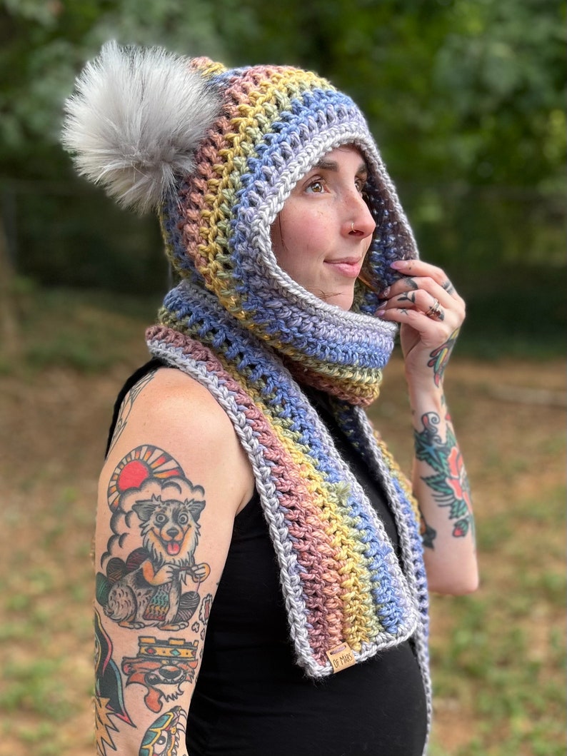 May include: A rainbow crochet hooded scarf with a fluffy pom pom. The scarf is made with a variety of colors, including pink, purple, blue, green, and yellow. The scarf is tied around the neck of a person with tattoos on their arms. The scarf has a tag that says "Of Mars".