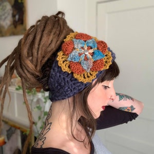 May include: A dark blue crochet headband featuring a large, multi-layered floral applique. The flower consists of rust-orange, light blue, and mustard-yellow petals with a small silver-toned circular button at the center of the bloom.