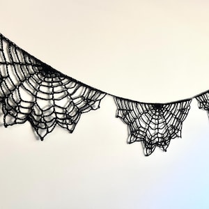 Op de afbeelding: Een zwarte, gehaakte spinnenweb slinger, ideaal voor Halloween-decoraties. De slinger heeft meerdere websecties van verschillende groottes, elk met ingewikkelde details. De spinnenwebben zijn opgehangen aan een dun zwart touwtje.
