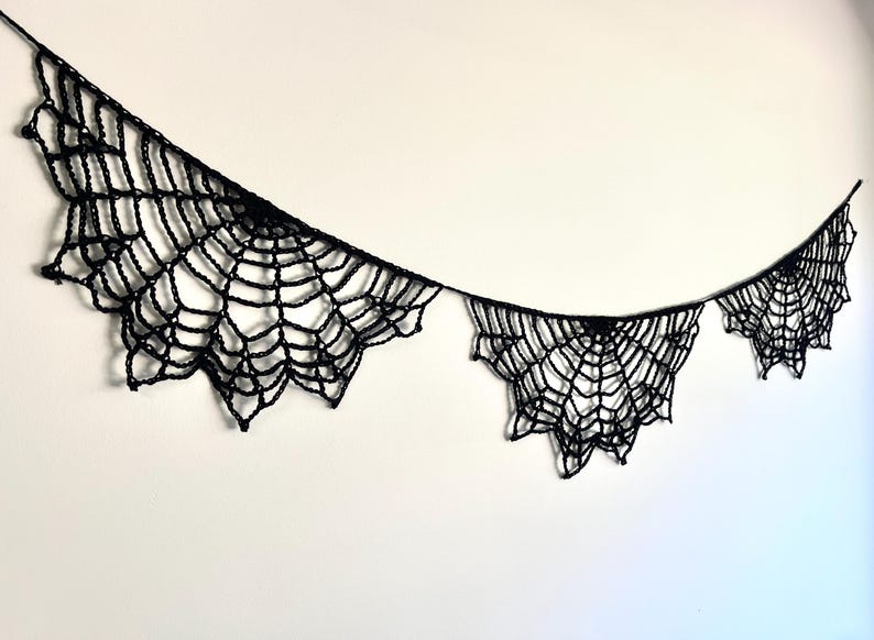 Op de afbeelding: Een zwarte, gehaakte spinnenweb slinger, ideaal voor Halloween-decoraties. De slinger heeft meerdere websecties van verschillende groottes, elk met ingewikkelde details. De spinnenwebben zijn opgehangen aan een dun zwart touwtje.
