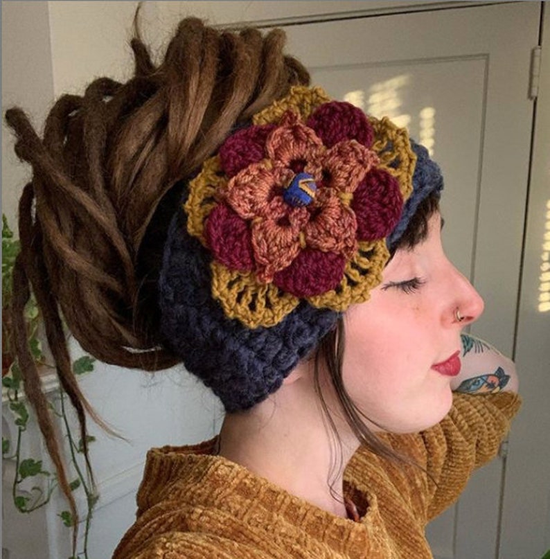May include: A dark blue crochet headband featuring a large, multi-layered floral applique. The flower consists of mustard yellow, burnt orange, and deep burgundy petals, with a small blue and yellow patterned button at the center of the bloom.