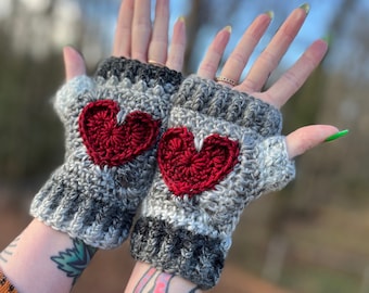 May include: A pair of gray and white fingerless gloves with red crocheted hearts. The gloves are made of a soft, fuzzy yarn.