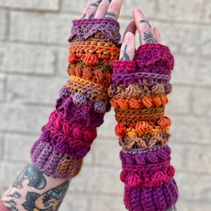 May include: A pair of colorful crocheted fingerless gloves with a multi-colored design. The gloves are made with a variety of stitches and textures, including bobbles and cables. The gloves are a vibrant mix of purple, orange, brown, and red.