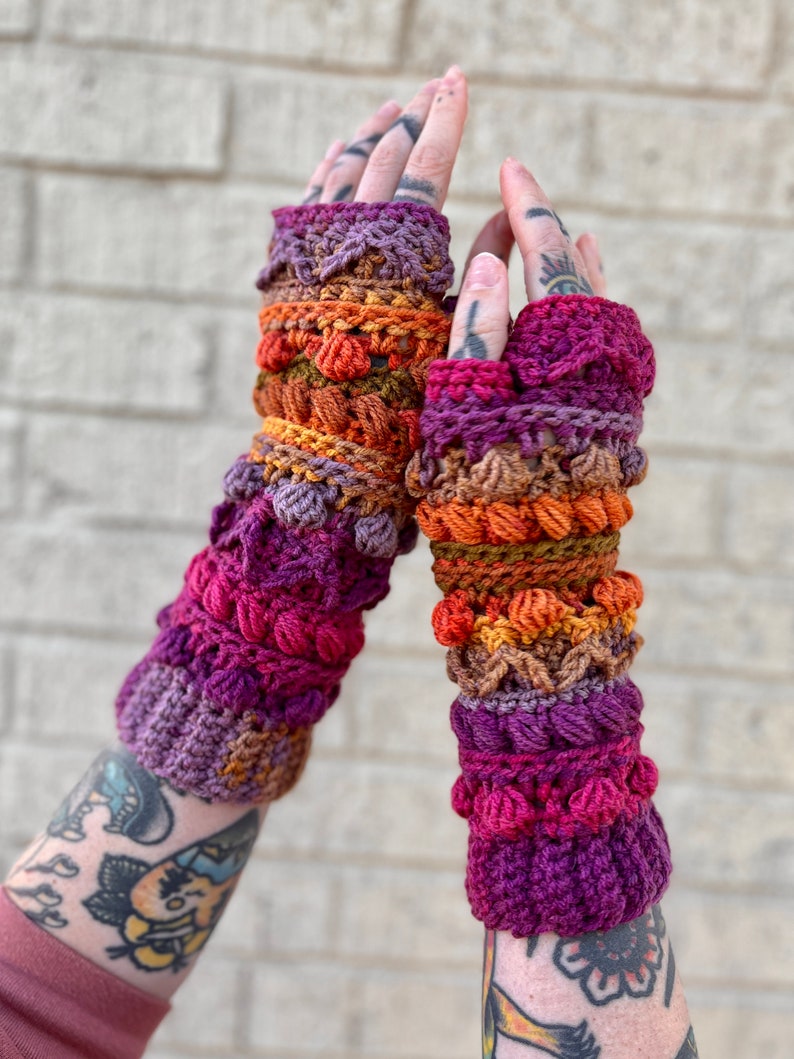May include: A pair of colorful crocheted fingerless gloves with a multi-colored design. The gloves are made with a variety of stitches and textures, including bobbles and cables. The gloves are a vibrant mix of purple, orange, brown, and red.