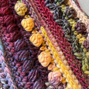 Op de afbeelding: Close-up van een handgemaakt gehaakt item met een verscheidenheid aan texturen en kleuren. Het stuk heeft verhoogde, getextureerde bobbles in tinten geel, roze en groen, tegen een achtergrond van diep rood, paars en blauw.