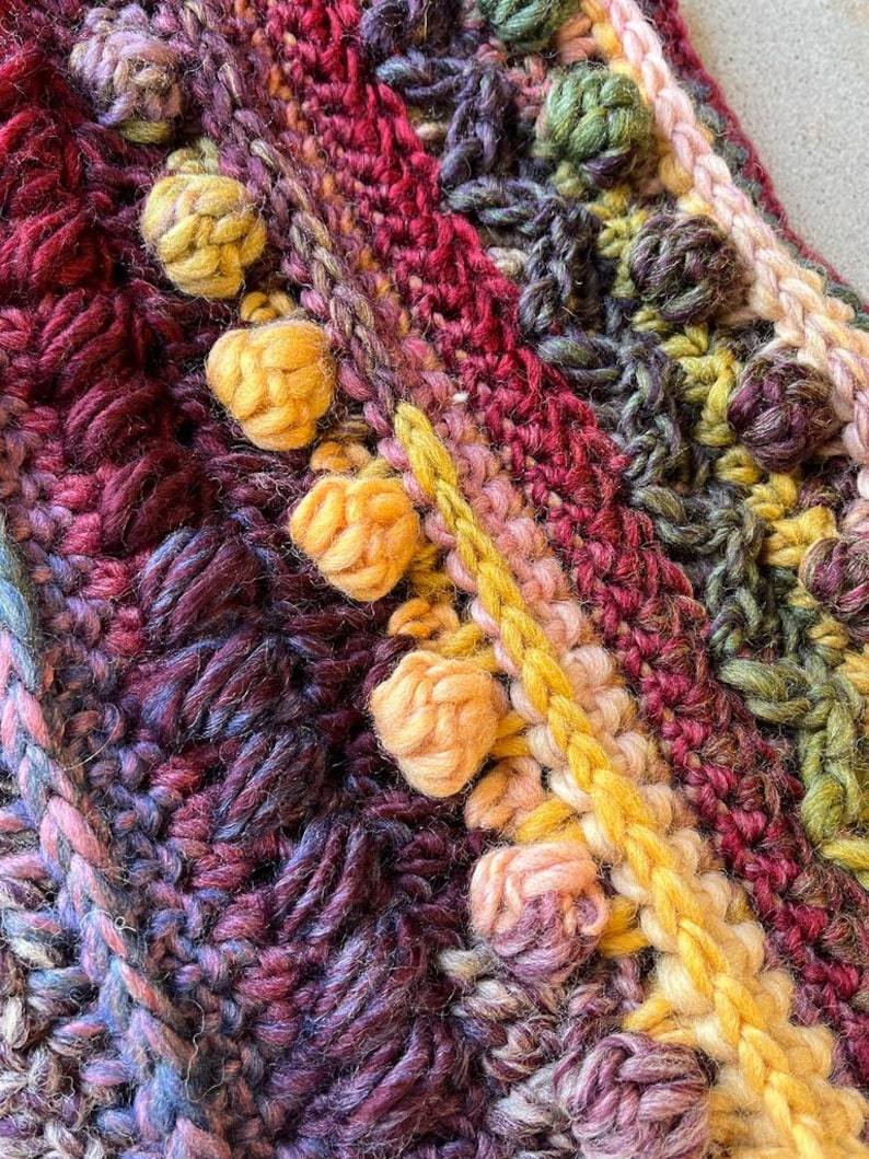 Op de afbeelding: Close-up van een handgemaakt gehaakt item met een verscheidenheid aan texturen en kleuren. Het stuk heeft verhoogde, getextureerde bobbles in tinten geel, roze en groen, tegen een achtergrond van diep rood, paars en blauw.