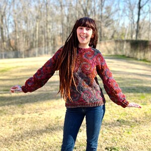 May include: A woman with long brown dreadlocks is wearing a red, orange and brown jumper with a circular pattern. She is smiling and wearing blue jeans.