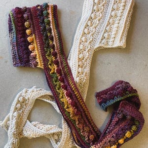 Op de afbeelding: Twee handgemaakte gehaakte sjaals. E&eacute;n is cr&egrave;mekleurig met een textuurpatroon. De andere is een levendige mix van kleuren, waaronder paars, groen en geel, met een textuur, bobbelachtig ontwerp. De sjaals liggen op een grijze ondergrond.
