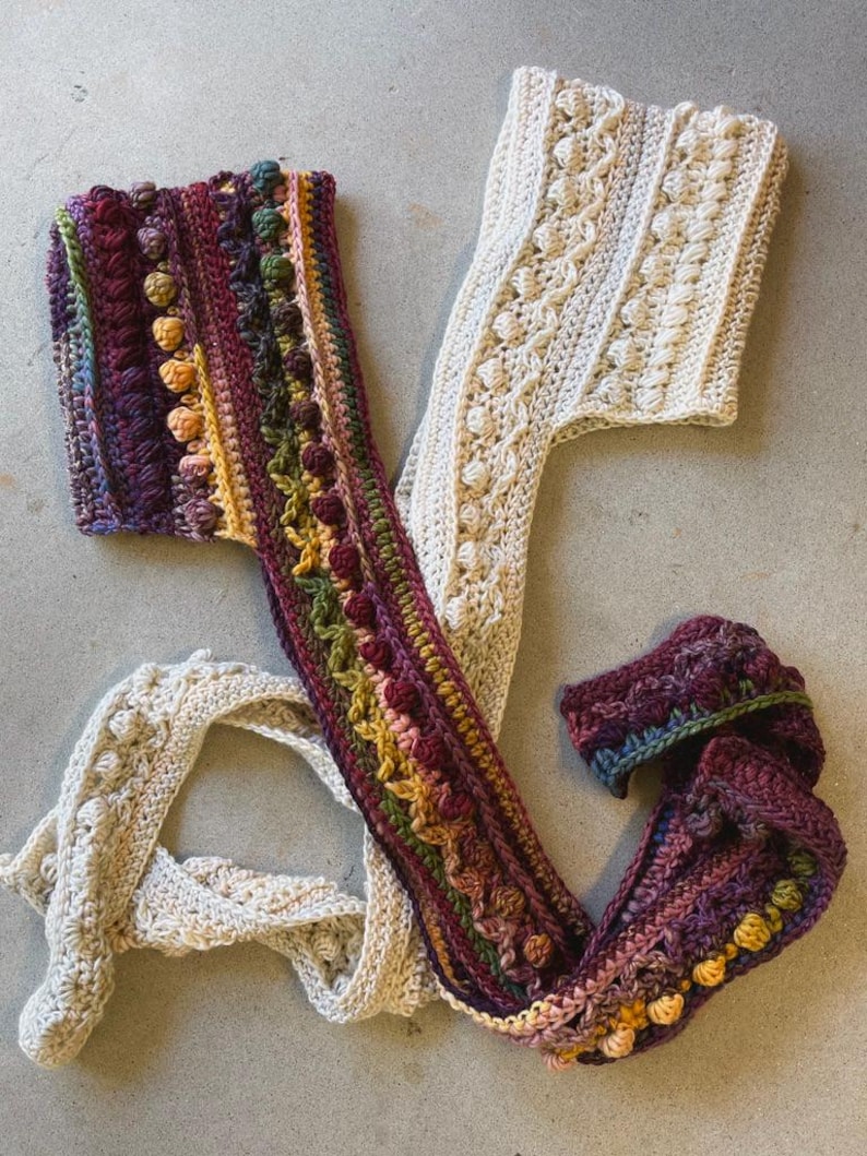 Op de afbeelding: Twee handgemaakte gehaakte sjaals. E&eacute;n is cr&egrave;mekleurig met een textuurpatroon. De andere is een levendige mix van kleuren, waaronder paars, groen en geel, met een textuur, bobbelachtig ontwerp. De sjaals liggen op een grijze ondergrond.