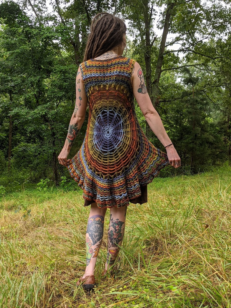 May include: A sleeveless crochet tunic featuring a circular spiderweb pattern in shades of orange, yellow, blue, and brown. The garment has a flared, ruffled hemline and an open-knit construction, layered over a solid brown base garment.