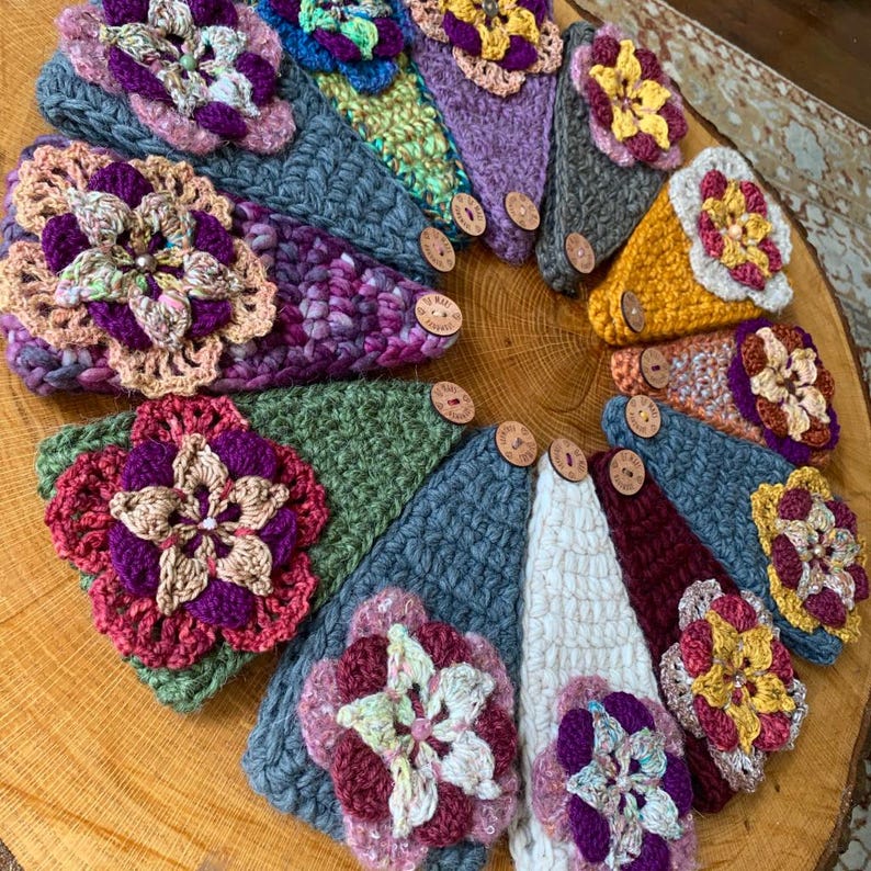 May include: A collection of triangular crochet fabric pieces arranged in a circle. Each piece features a textured knit pattern in various colors including gray, green, white, and purple, adorned with a large, multi-layered crochet flower.