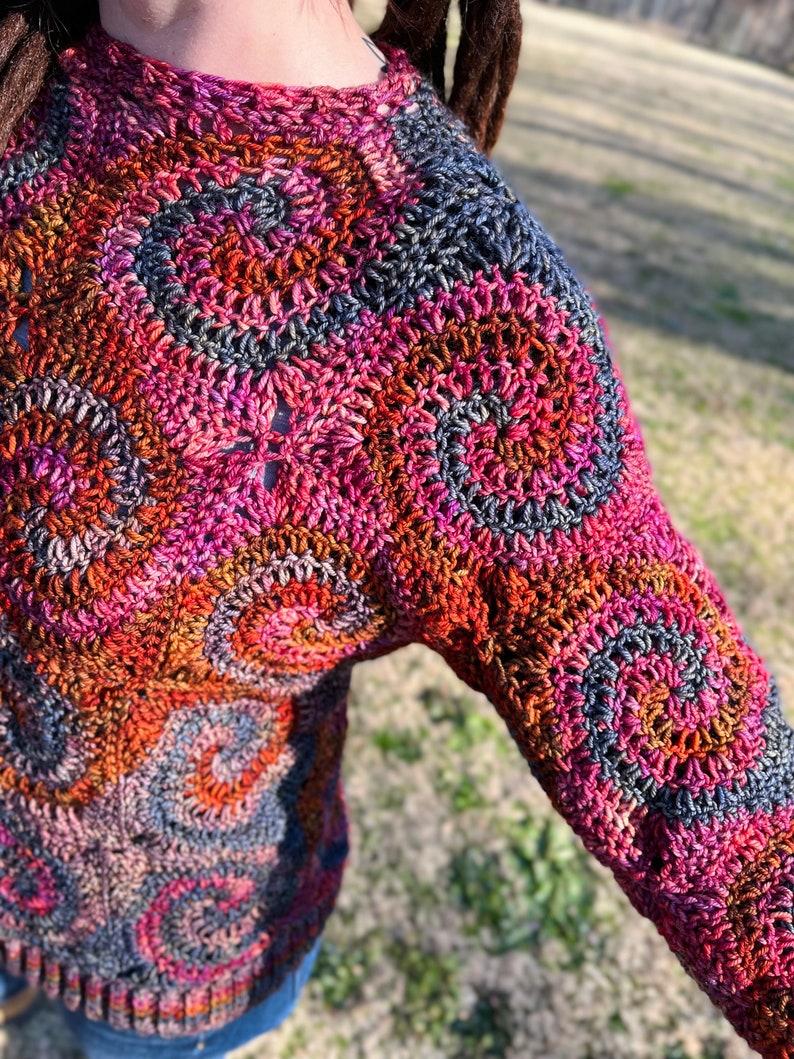 May include: A colourful crochet sweater with a circular pattern in shades of pink, orange, brown, and grey.