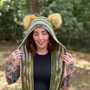 May include: A green, white, and brown striped hooded scarf with faux fur earmuffs. The scarf is crocheted and has a long, flowing design.