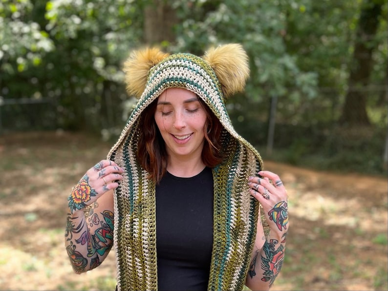 May include: A green, white, and brown striped hooded scarf with faux fur earmuffs. The scarf is crocheted and has a long, flowing design.