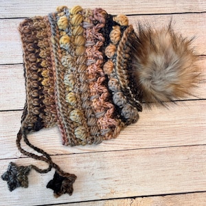 May include: A hand-knitted hat in shades of brown, beige, and pink, with a faux fur pom-pom and star-shaped tassels. The hat's textured design is visible, and the pom-pom is a light brown colour. The tassels are star-shaped.