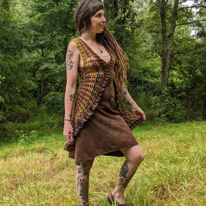 May include: A sleeveless, open-front crochet vest featuring a multi-colored pattern of orange, yellow, purple, and blue yarn. The vest has a V-neckline and a lace-up front closure with thin ties. It is worn over a solid brown, knee-length dress.