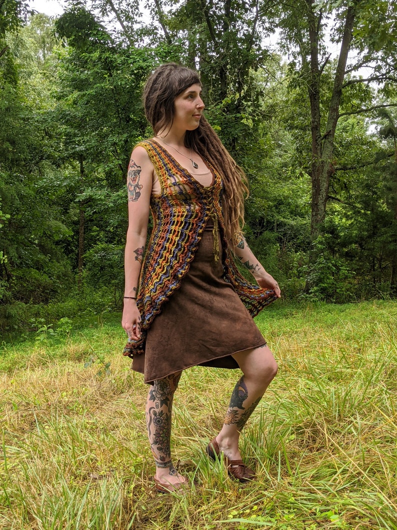 May include: A sleeveless, open-front crochet vest featuring a multi-colored pattern of orange, yellow, purple, and blue yarn. The vest has a V-neckline and a lace-up front closure with thin ties. It is worn over a solid brown, knee-length dress.