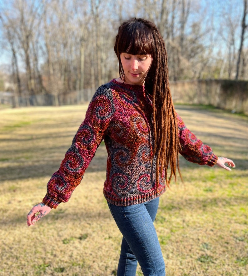 May include: A colourful crocheted cardigan with a textured pattern in shades of red, brown, and blue. The cardigan has a relaxed fit and a slightly cropped length.
