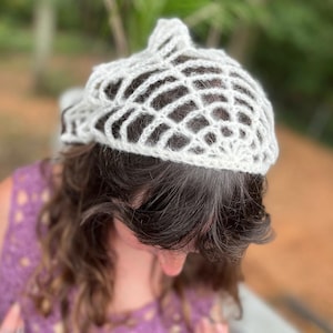 May include: A white, open-weave crochet hat. The hat has a rounded crown and a delicate, web-like pattern. The hat is made of a soft, light-colored yarn. The hat is worn on a person's head, with their dark hair visible beneath.