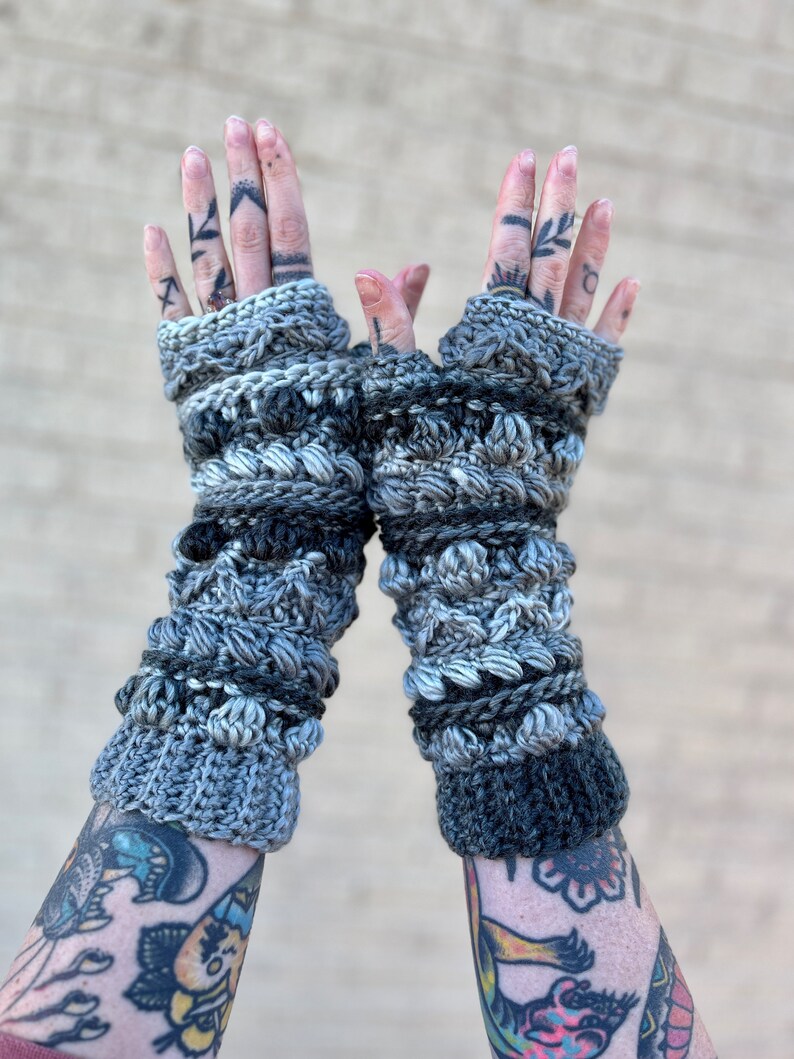 May include: A pair of gray and black fingerless crochet gloves with a textured design. The gloves are made with a variety of stitches and yarns, creating a unique and stylish look.