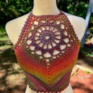 May include: A crocheted halter top with a colorful ombre design in shades of red, orange, yellow, green, and purple. The top features a large crocheted flower design at the neckline.