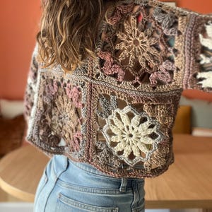 May include: A crocheted top in shades of brown, beige and pink. The top is made using a variety of stitches and patterns, including granny squares and flowers. The top is cropped and has long sleeves.