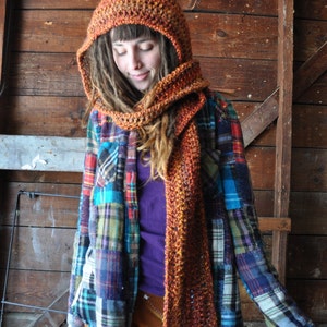 May include: A woman wearing a brown and orange crocheted hooded scarf and matching hat. She is also wearing a plaid shirt and brown pants.