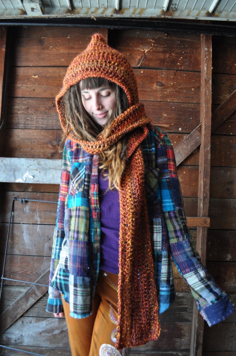 May include: A woman wearing a brown and orange crocheted hooded scarf and matching hat. She is also wearing a plaid shirt and brown pants.