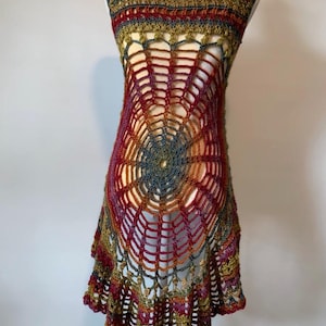 May include: A sleeveless crochet tunic featuring a prominent spiderweb pattern in the center. The garment is crafted from variegated yarn in shades of deep red, olive green, and muted blue, with a flared, scalloped hemline and an open-work design.