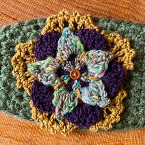 May include: A layered crochet flower applique featuring a dark green base, a mustard yellow scalloped layer, deep purple petals, and a central variegated green, blue, and white yarn flower with a small brown button at the very center.