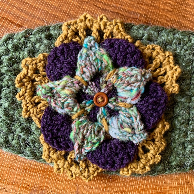 May include: A layered crochet flower applique featuring a dark green base, a mustard yellow scalloped layer, deep purple petals, and a central variegated green, blue, and white yarn flower with a small brown button at the very center.