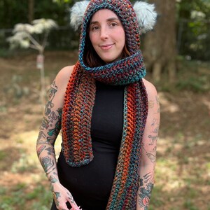 May include: A woman wearing a black dress with a multi-colored crocheted scarf and matching hooded hat with faux fur earmuffs.