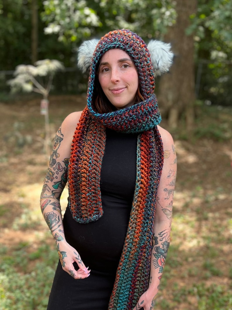 May include: A woman wearing a black dress with a multi-colored crocheted scarf and matching hooded hat with faux fur earmuffs.