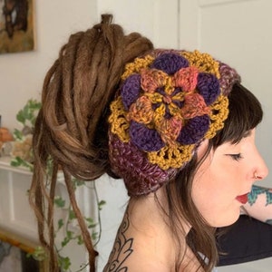 May include: A crochet headband featuring a large, multi-layered floral motif in shades of mustard yellow, deep purple, and variegated orange. The accessory is constructed with a textured yarn pattern, showcasing a wide band design that wraps around the head.