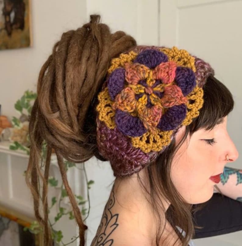 May include: A crochet headband featuring a large, multi-layered floral motif in shades of mustard yellow, deep purple, and variegated orange. The accessory is constructed with a textured yarn pattern, showcasing a wide band design that wraps around the head.
