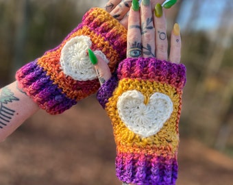 May include: A pair of fingerless gloves with a crocheted heart design. The gloves are made with a rainbow of colors, including purple, orange, yellow, and white.