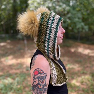 May include: A green and beige striped crocheted hooded scarf with two fluffy pom-poms. The scarf is worn by a person with a tattoo on their arm.
