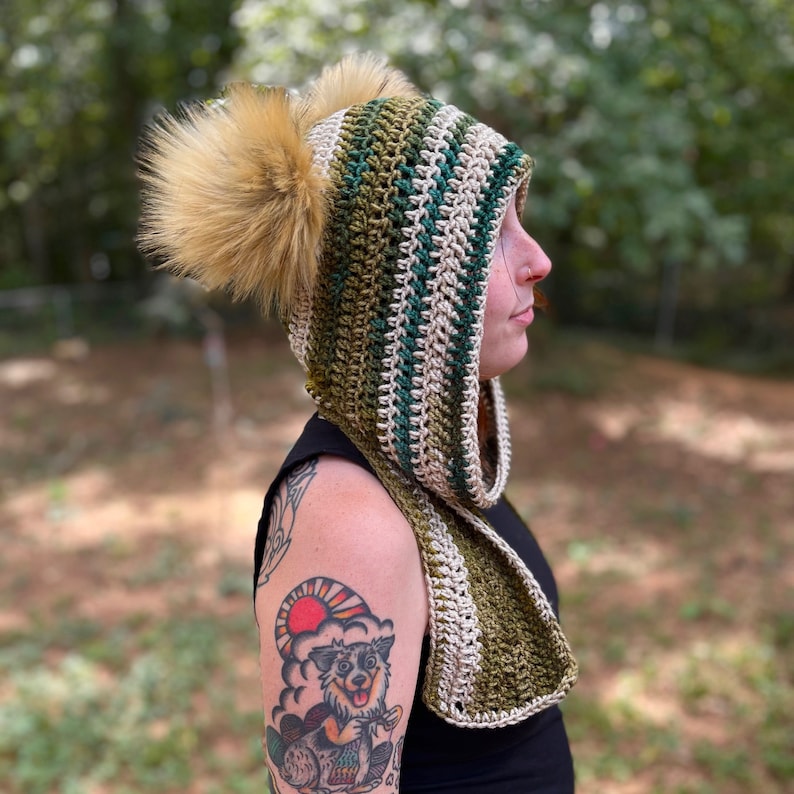 May include: A green and beige striped crocheted hooded scarf with two fluffy pom-poms. The scarf is worn by a person with a tattoo on their arm.