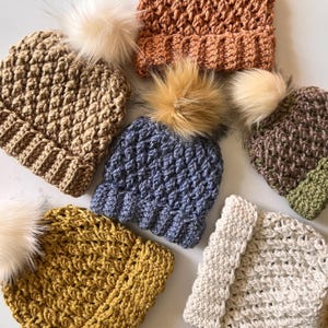 May include: Six crocheted hats in various colors, including beige, brown, blue, yellow, and white. Each hat has a fluffy pom-pom on top.