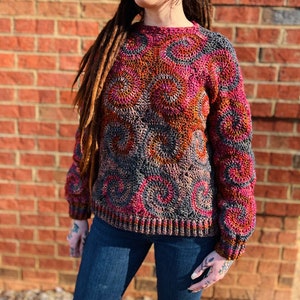 May include: A crocheted sweater with a colourful, swirling pattern in shades of pink, orange, brown, and grey. The sweater has a round neckline and long sleeves.