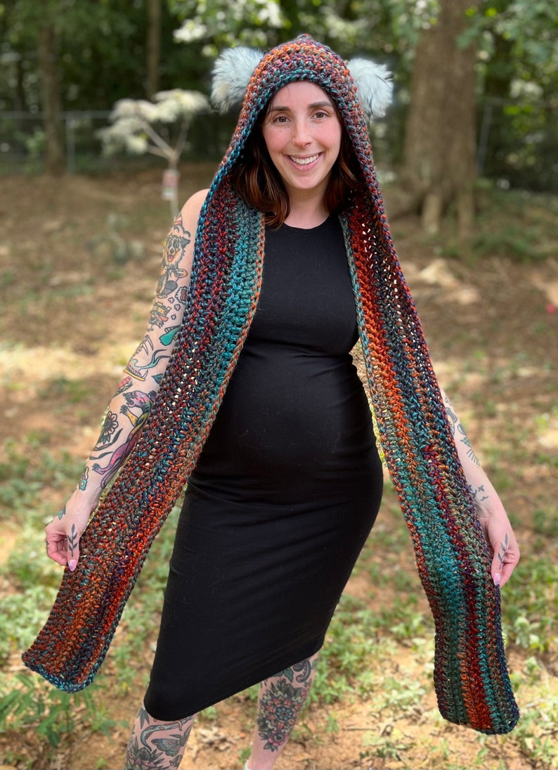 May include: A crocheted hooded scarf with a multi-colored striped pattern in shades of orange, teal, and brown. The scarf has a hood with two fluffy white ear-like accents.