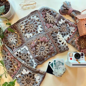 K&ouml;nnte beinhalten: Eine geh&auml;kelte Strickjacke mit einem Granny-Square-Muster in Braun-, Rosa- und Wei&szlig;t&ouml;nen. Die Strickjacke liegt flach auf einer wei&szlig;en Oberfl&auml;che mit einer Vintage-Kamera und anderem Zubeh&ouml;r.