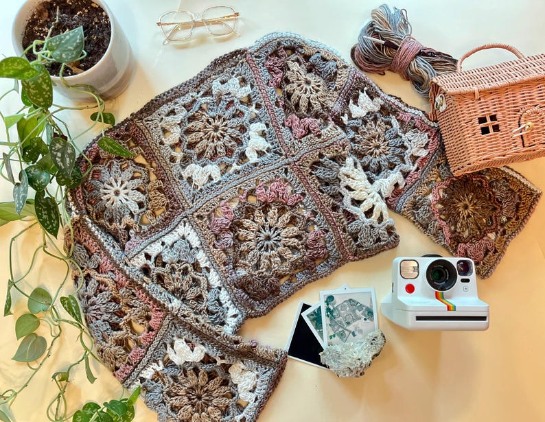 K&ouml;nnte beinhalten: Eine geh&auml;kelte Strickjacke mit einem Granny-Square-Muster in Braun-, Rosa- und Wei&szlig;t&ouml;nen. Die Strickjacke liegt flach auf einer wei&szlig;en Oberfl&auml;che mit einer Vintage-Kamera und anderem Zubeh&ouml;r.