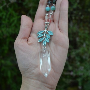 May include: A clear quartz crystal pendant necklace with a silver-coloured leaf charm and colourful beaded accents. The pendant is suspended from a chain with turquoise, pink, and brown beads. The leaf charm has a teal finish.