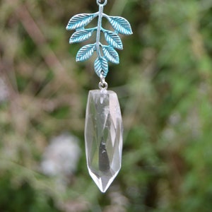 May include: A clear quartz crystal pendant with a faceted design hangs from a beaded chain. The pendant is suspended from a teal-coloured metal leaf charm. The chain features pink and white beads.
