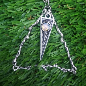 May include: A silver necklace with a triangular pendant featuring a small, iridescent gemstone. The pendant is attached to a delicate chain and hangs from a simple, open triangle frame.