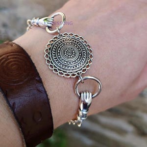 May include: A silver-toned bracelet featuring a central circular medallion with intricate geometric patterns and scalloped edges. The chain is composed of links shaped like small hands holding rings, with the text VALKYRIESSONG visible.
