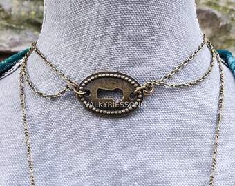 Puede incluir: Un collar dorado con un colgante en forma de ojo de cerradura. El colgante tiene la inscripción "VALKYRIESSONG".