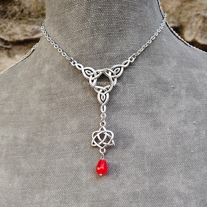 Può includere: Collana con pendente a nodo celtico color argento con una perla rossa a forma di goccia. La collana presenta intricati disegni a nodo ed è esposta su un manichino grigio. La catena è delicata.